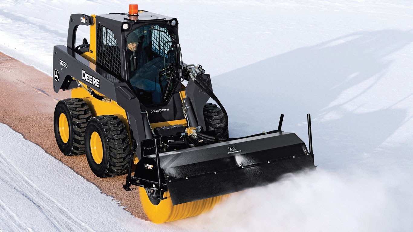 Une chargeuse à direction différentielle John Deere équipée de l’accessoire balai orientable balaie la neige.